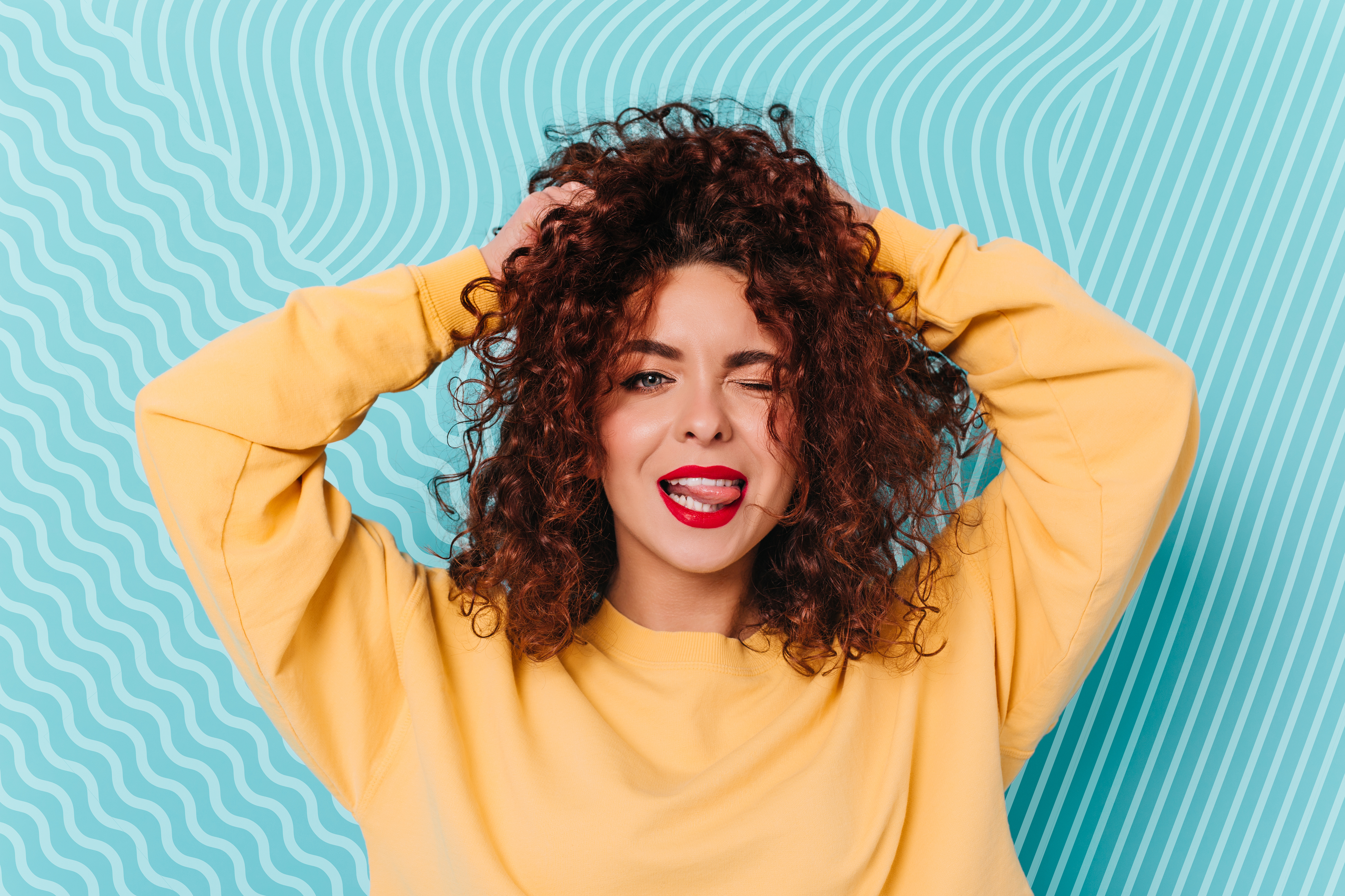 A woman wearing a yellow sweatshirt playfully runs her hands through her curly hair, winking and sticking out her tongue.